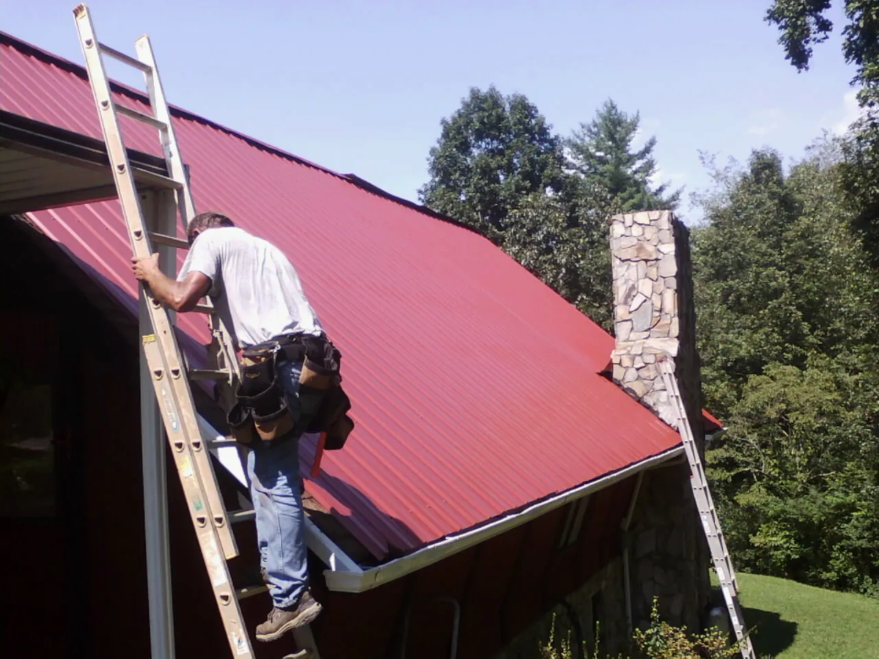 Roofer on a ladder beside a red metal roof and stone chimney — Soffit & Fascia Repair by Roofing Brothers in Running Springs